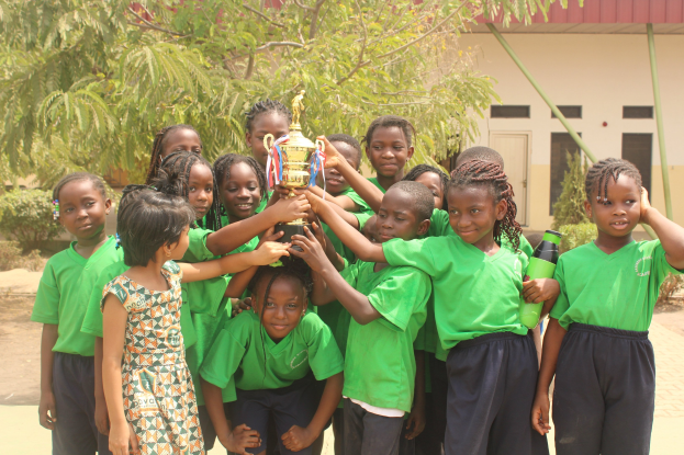 Eine Gruppe von Kindern, die einen Pokal in einem Schulhof mit Bäumen, Pflanzen und einem Gebäude im Hintergrund halten.