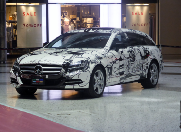 Ein silberner Mercedes-Benz CLS Shooting Brake vor einem Geschäft mit Schaufensterpuppen in Kleidern im Hintergrund, eine Säule links und eine Wand rechts.