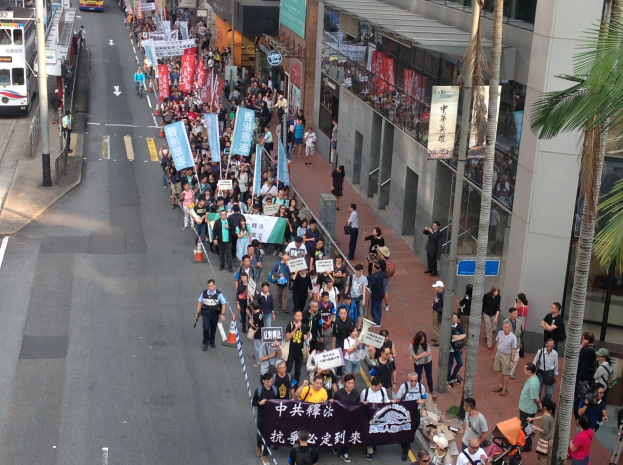 Eine große Gruppe von Menschen marschiert mit Protestschildern und Transparenten eine Straße entlang, mit Bäumen und gläsernen Gebäuden auf der rechten Seite, Fahrzeugen auf der linken Seite und Straßenschildern und Pfählen im Bild.
