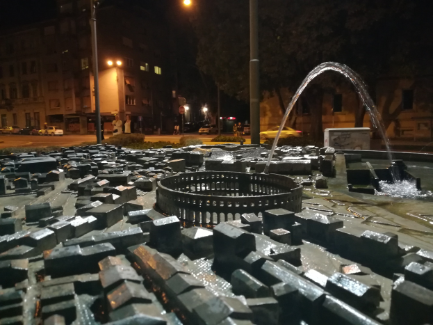 Ein nächtlicher Stadtplatz mit einem zentralen Brunnen, umgeben von Straßenlaternen, Bäumen, Gebäuden mit beleuchteten Fenstern, Fahrzeugen auf der Straße und einigen fernen Passanten unter einem dunklen Himmel.