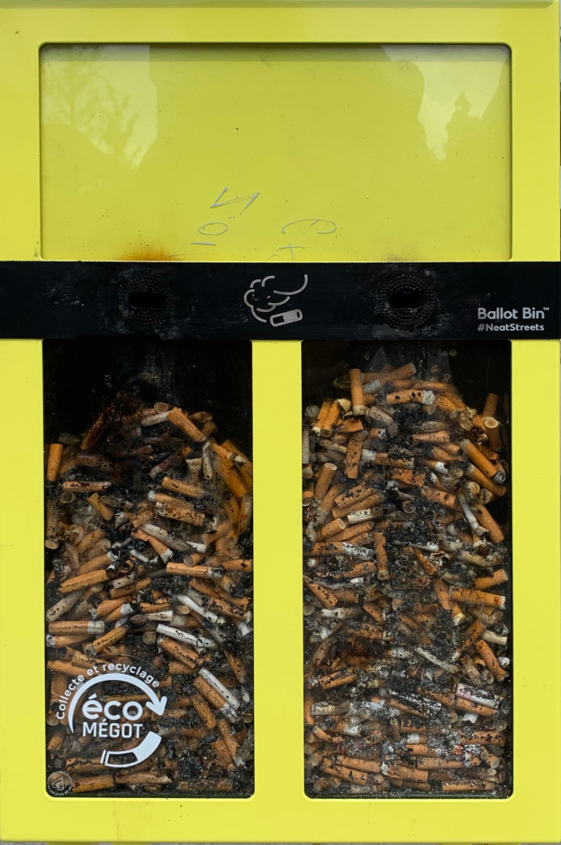 Ein leuchtend gelber Recyclingmüllbehälter voller Zigarettenstummel auf einem Gehweg, beschriftet für die Entsorgung von Zigarettenabfall.