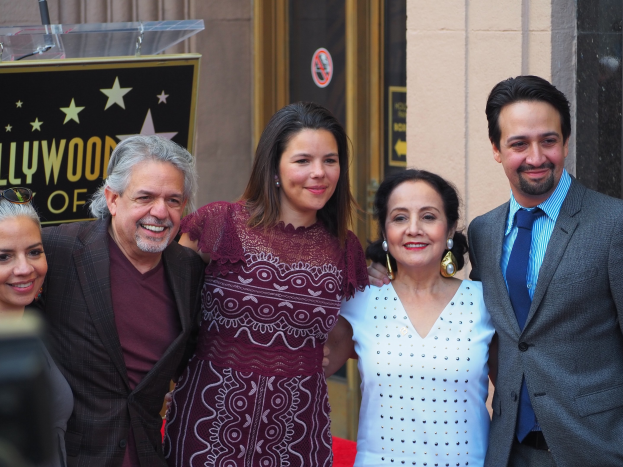 Gruppe von Menschen, die auf einem roten Teppich an der Hollywood Walk of Fame Zeremonie posen, mit einer Kamera auf der linken Seite, einer Tafel mit Text, einem Glaspodest und einer Wand im Hintergrund.