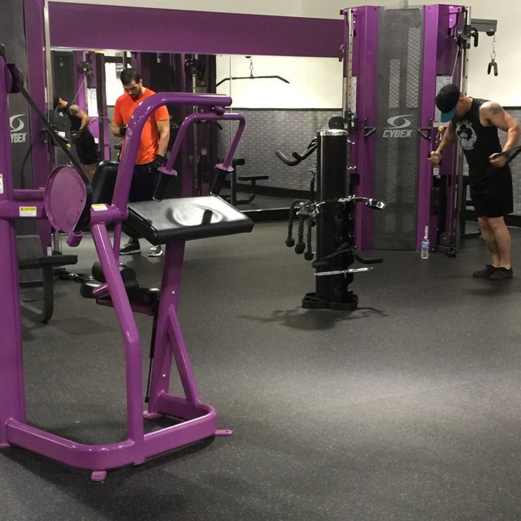 Eine Person in einem schwarzen Kleid, einer Mütze und Schuhen steht in einem Fitnessstudio mit verschiedenen Geräten, während eine andere Person in einem orangen T-Shirt und Schuhen in der Nähe steht, wobei ein Spiegel im Hintergrund zu sehen ist.