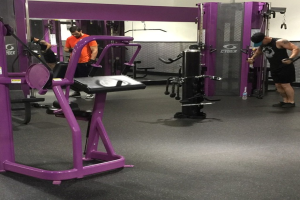 Eine Person in einem schwarzen Kleid, einer Mütze und Schuhen steht in einem Fitnessstudio mit verschiedenen Geräten, während eine andere Person in einem orangen T-Shirt und Schuhen in der Nähe steht, wobei ein Spiegel im Hintergrund zu sehen ist.