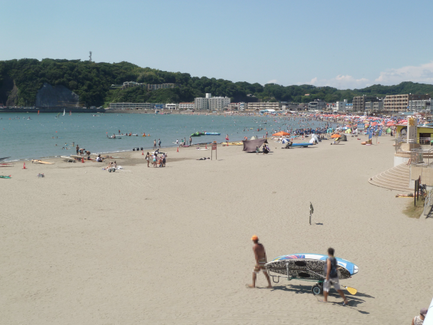 Ein voller Strand mit Menschen, einige halten Surfbretter, Sonnenschirme, Zelte und Treppen zum Wasser, mit Gebäuden, Bäumen, Hügeln und einem bewölkten Himmel im Hintergrund.