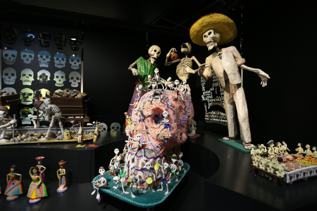 Eine Ausstellung zum Tag der Toten im Fine Arts Museum in San Francisco, die Spielzeug, Skelette und andere Gegenstände auf Tischen zeigt, mit einer Texttafel im Hintergrund, in einer dunklen und mysteriösen Atmosphäre.