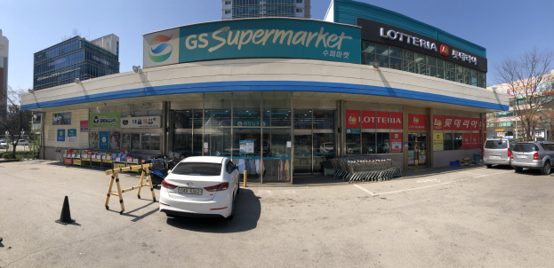 Ein Auto vor einem "GS Supermarkt"-Geschäft mit anderen Fahrzeugen, Verkehrskegeln und verstreuten Gegenständen auf der Straße; Gebäude, Bäume und ein klarer blauer Himmel im Hintergrund.