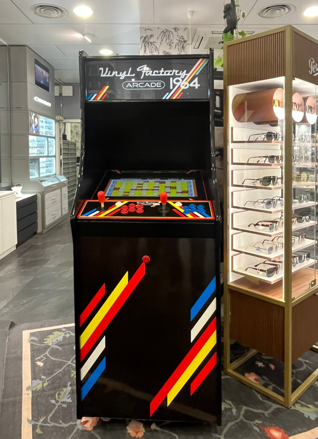 Ein Geschäft mit einem zentralen Retro-Arcade-Automaten, einem Gestell mit Gläsern auf der rechten Seite, Schränken und einem Fernseher im Hintergrund und einer Deckenbeleuchtung, die eine retro-mäßige Atmosphäre schafft.