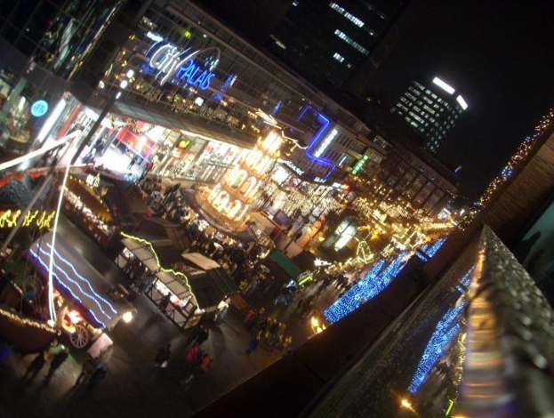 Eine belebte Stadtstraße bei Nacht, voller Menschen, die unter der Beleuchtung von Gebäuden und Ständen gehen, mit einem dunklen Himmel im Hintergrund.