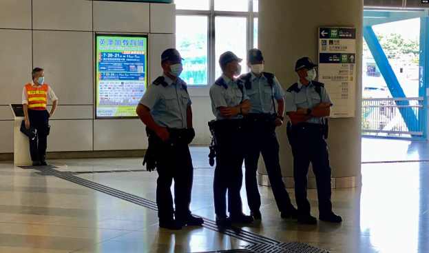 Eine Gruppe maskierter Polizisten steht vor einem Gebäude mit Glastüren, Schildern, einer Säule und einem Geländer, mit Bäumen und Fahrzeugen im Hintergrund, was darauf hinweist, dass sie in Hong Kong festgenommen wurden.