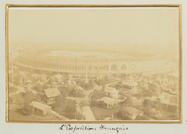 Altes Foto eines Stadions in einer städtischen Umgebung mit umliegenden Gebäuden, Bäumen und Masten, mit Text unten.
