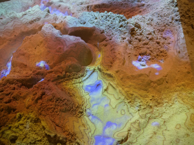 Nahaufnahme einer detaillierten 3D-topografischen Karte einer Wüstenlandschaft, die Berge, Täler und Grate mit Sandfarben von hell bis dunkelbraun zeigt, die von hellem Licht beleuchtet werden.