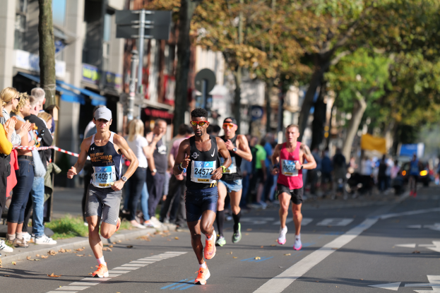 Eine Gruppe von Menschen, die bei einem Marathon auf einer Straße laufen, mit Zuschauern auf der linken Seite, Bäumen, Gebäuden und einem klaren blauen Himmel im Hintergrund, alle Läufer tragen Turnschuhe und Hüte.