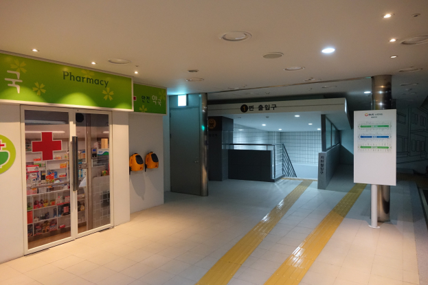 Apotheken-Eingang mit einem 'Apotheke'-Schild auf der linken Seite, einer Tafel mit Text auf der rechten Seite, beleuchteten Lampen oben und Hintergrundobjekten.