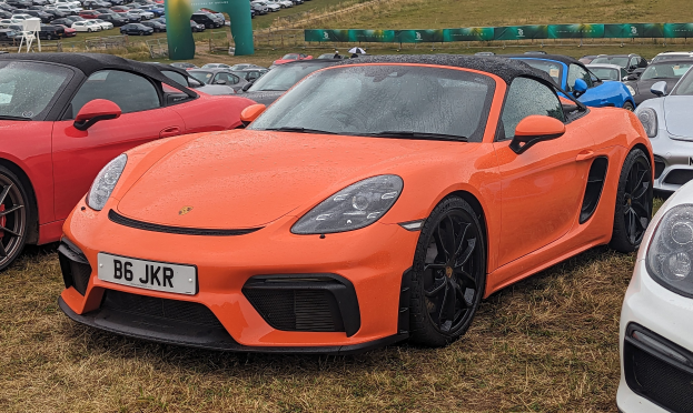 Roter Porsche 718 Boxster GTS auf dem Goodwood Festival of Speed, umgeben von anderen Autos auf dem Rasen, mit einem Banner, Bäumen und einem klaren blauen Himmel im Hintergrund.