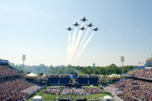 Eine Gruppe von Jets fliegt niedrig über ein Footballfeld mit Zuschauern in den Rängen, Bäumen, Gebäuden und einem klaren blauen Himmel im Hintergrund.