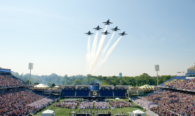 Eine Gruppe von Jets fliegt niedrig über ein Footballfeld mit Zuschauern in den Rängen, Bäumen, Gebäuden und einem klaren blauen Himmel im Hintergrund.