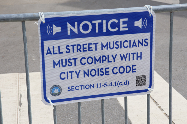 Anschlagtafel mit einem Schild, auf dem "Hinweis: Alle Straßenmusiker müssen die Stadtlärmvorschriften einhalten" steht, an einem Zaun befestigt, mit einer Straße im Hintergrund.