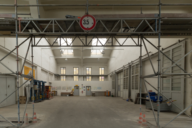 Ein großes Lagerhaus mit ausgedehnten Gerüsten, Bodenmarkierungen, Metallstangen, ein Schild, verschiedene Maschinen, eine Tür, Fenster, eine Wand und ein Dach mit Deckenleuchten.