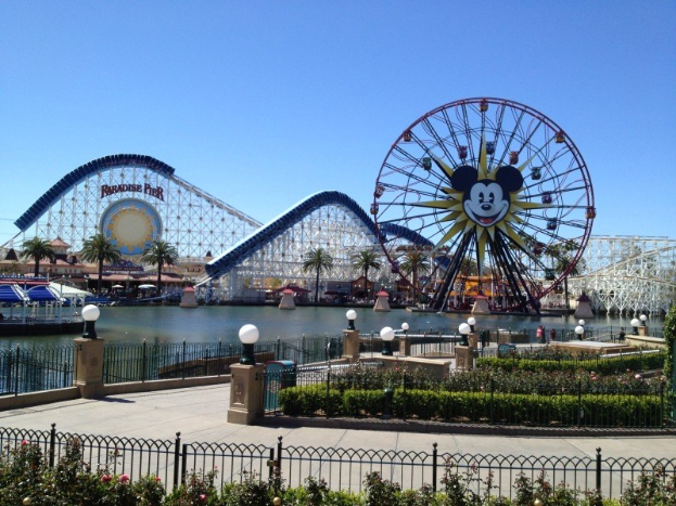 Luftaufnahme von Disney California Adventure mit einem Riesenrad, einer Achterbahn, Grünflächen, Laternenmasten, einem Gewässer und einem hellblauen Himmel.