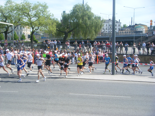 Gruppe von Menschen, die bei einem Marathon auf einer Straße mit Absperrungen, Zuschauern und städtischer Umgebung unter einem bewölkten Himmel laufen.