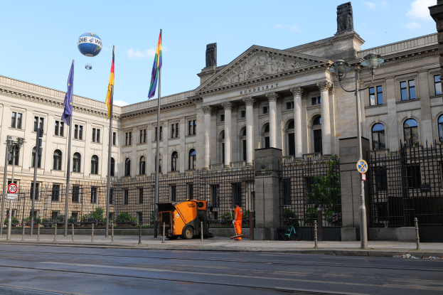 Das Reichstagsgebäude in Berlin, Deutschland, ein großes Gebäude mit zahlreichen Fenstern, umgeben von Straßenmasten, Fahrzeugen, Menschen und Bäumen, unter einem bewölkten Himmel.