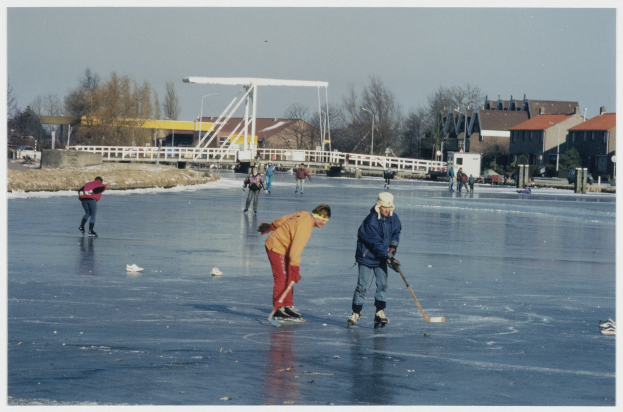 Eine Gruppe von Menschen, die Eis hockey auf einem zugefrorenen See spielt, mit Bäumen, Gebäuden, Pfählen und einer Brücke im Hintergrund, unter einem von Straßenlaternen beleuchteten Himmel.