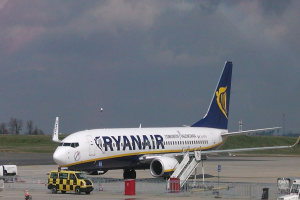 Ein Ryanair-Flugzeug auf dem Rollfeld eines Flughafens geparkt, umgeben von Fahrzeugen, Verkehrskegeln, Absperrungen, Pfählen, Schildern und anderen Gegenständen, mit Gras, Bäumen und einem bewölkten Himmel im Hintergrund.