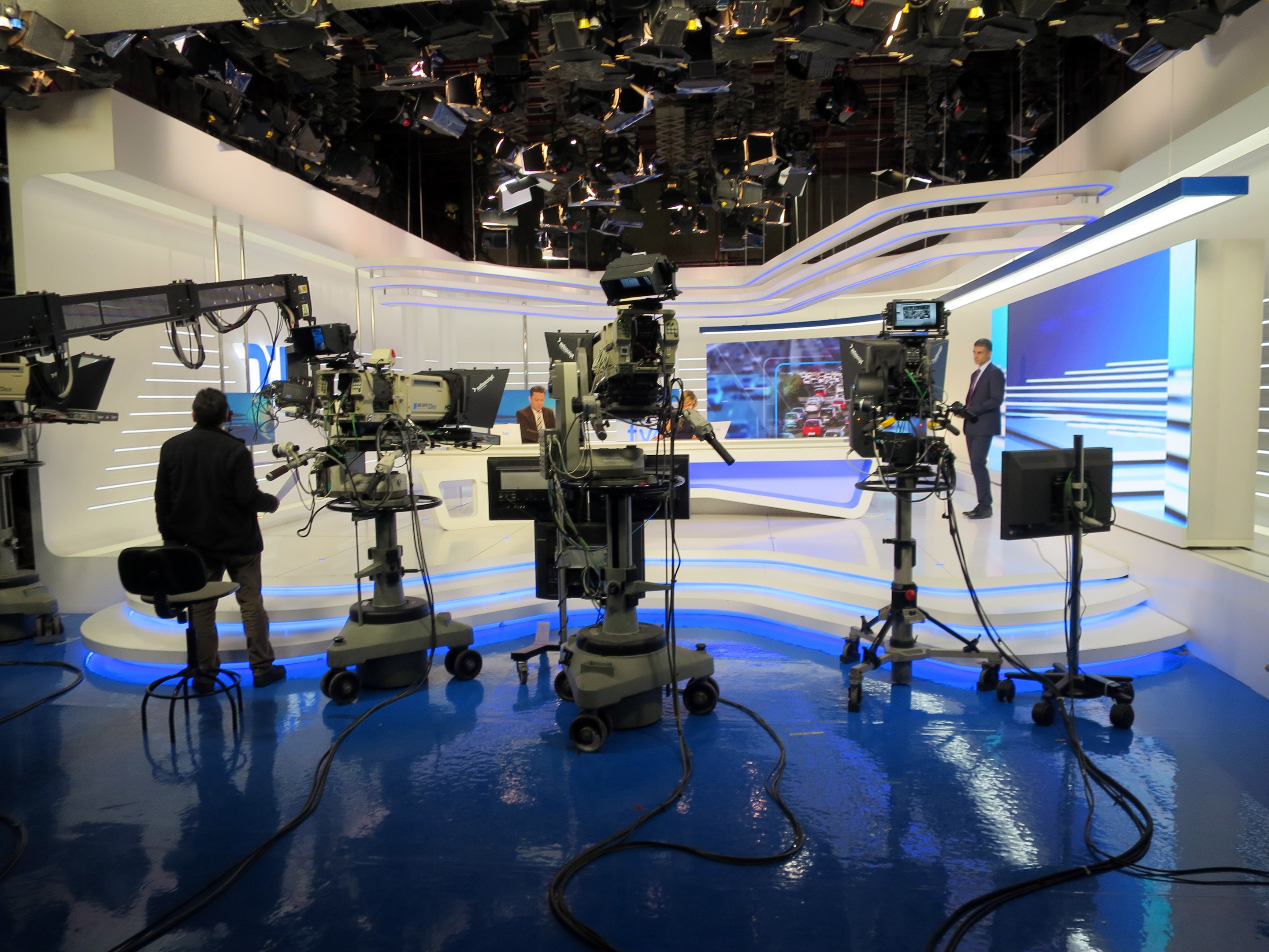 Ein Fernsehstudio mit mehreren Kameras auf dem Boden, einer sitzenden Person im Vordergrund und einigen stehenden Personen im Hintergrund, mit deckenmontierten Lampen für eine Livesendung.