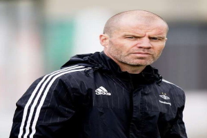 Ein kahlköpfiger Trainer in einer blauen Jacke mit weißen Streifen und weißem Kragen steht auf einem Fußballfeld mit verschränkten Händen und blickt ernst nach vorne.