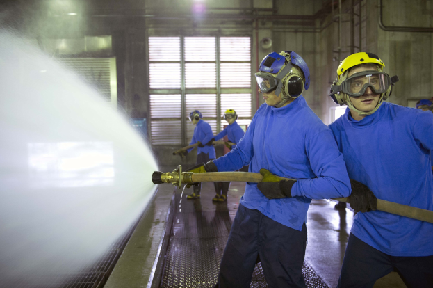 Gruppe von Männern in blauen Hemden und gelben Helmen bei der Arbeit an Maschinen in einer Fabrik mit sichtbaren Rohren, Fenstern und Lampen.