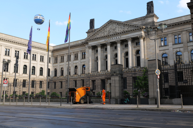 Das Bundestagsgebäude in Berlin, Deutschland, ein großes Gebäude mit zahlreichen Fenstern, umgeben von Straßenmasten, Fahrzeugen, Menschen und Bäumen unter einem bewölkten Himmel.